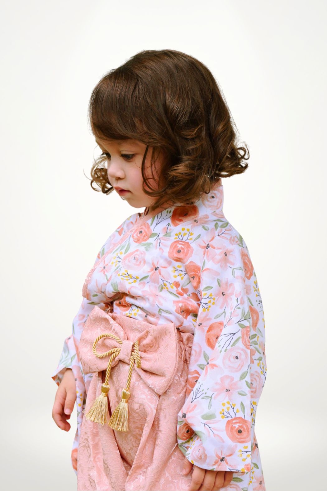 Une petite fille porte un yukata à fleurs roses avec un obi rose, se tenant de profil sur fond clair
