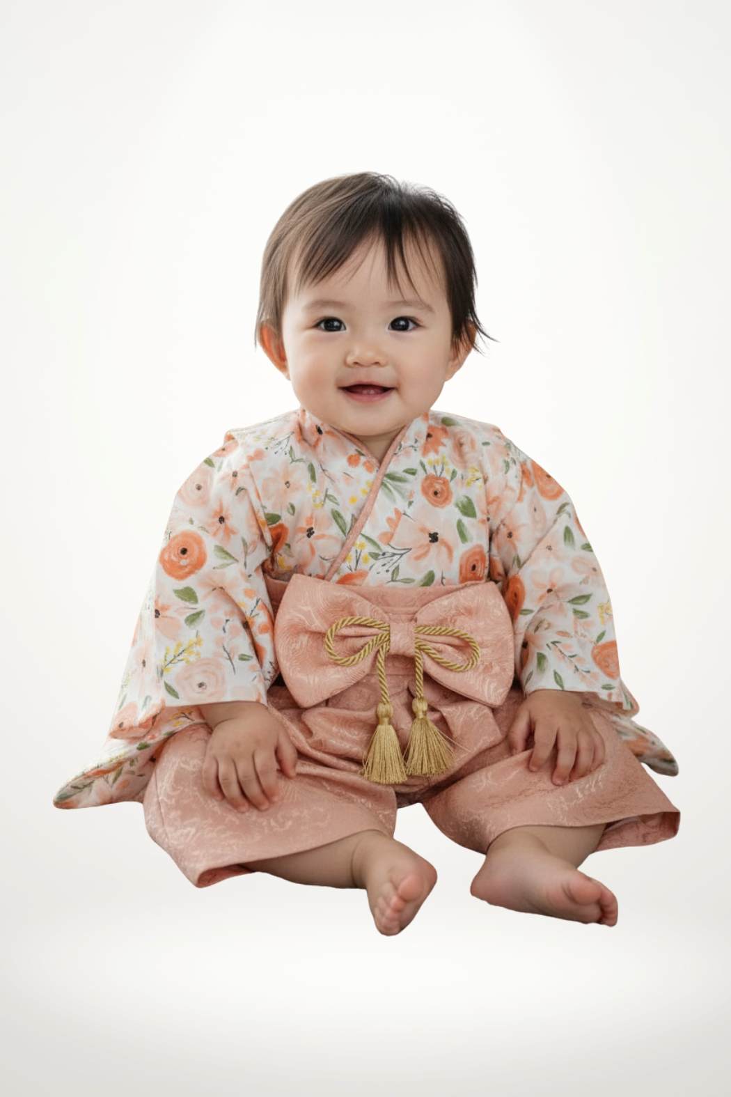 Un bébé souriant porte un yukata à fleurs roses et un pantalon en satin doré, assis sur un fond blanc