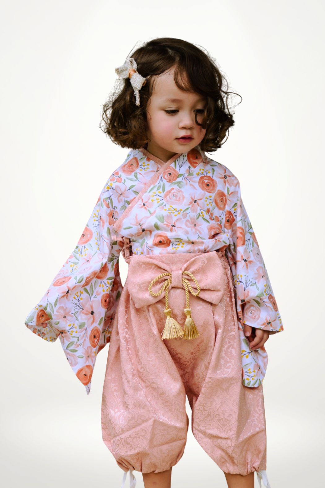 Une petite fille porte un yukata aux motifs de fleurs pêche, avec un pantalon rose et un décor minimaliste