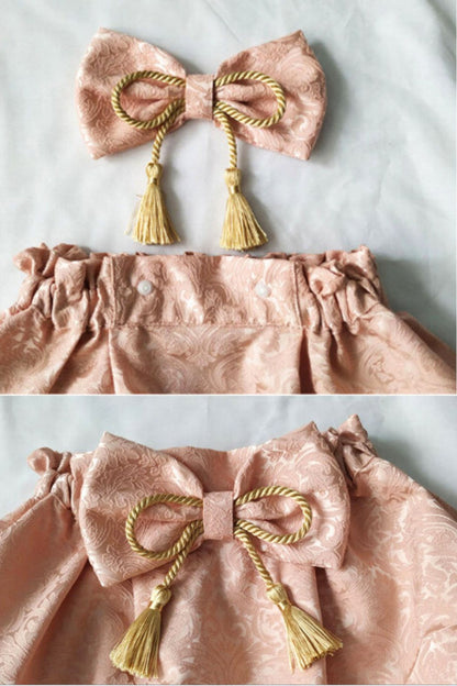 Un nœud en satin rose avec des franges dorées, sur un tissu à motifs délicats, évoquant l'élégance japonaise