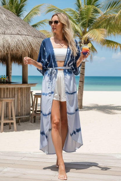 Femme en kimono bleu avec motifs tie-dye, portant un short blanc, se tenant sur une plage ensoleillée
