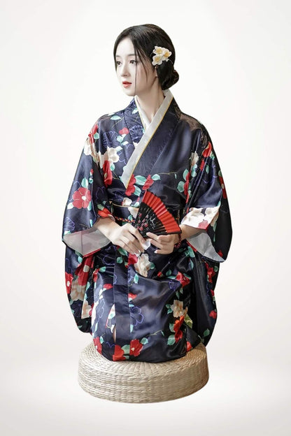 Femme assise en kimono satiné bleu marine orné de fleurs colorées, tenant un éventail rouge, sur un support en osier