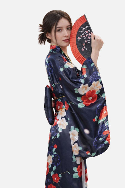 Femme en kimono satin bleu marine orné de fleurs colorées, tenant un éventail noir et rouge, dans une posture élégante