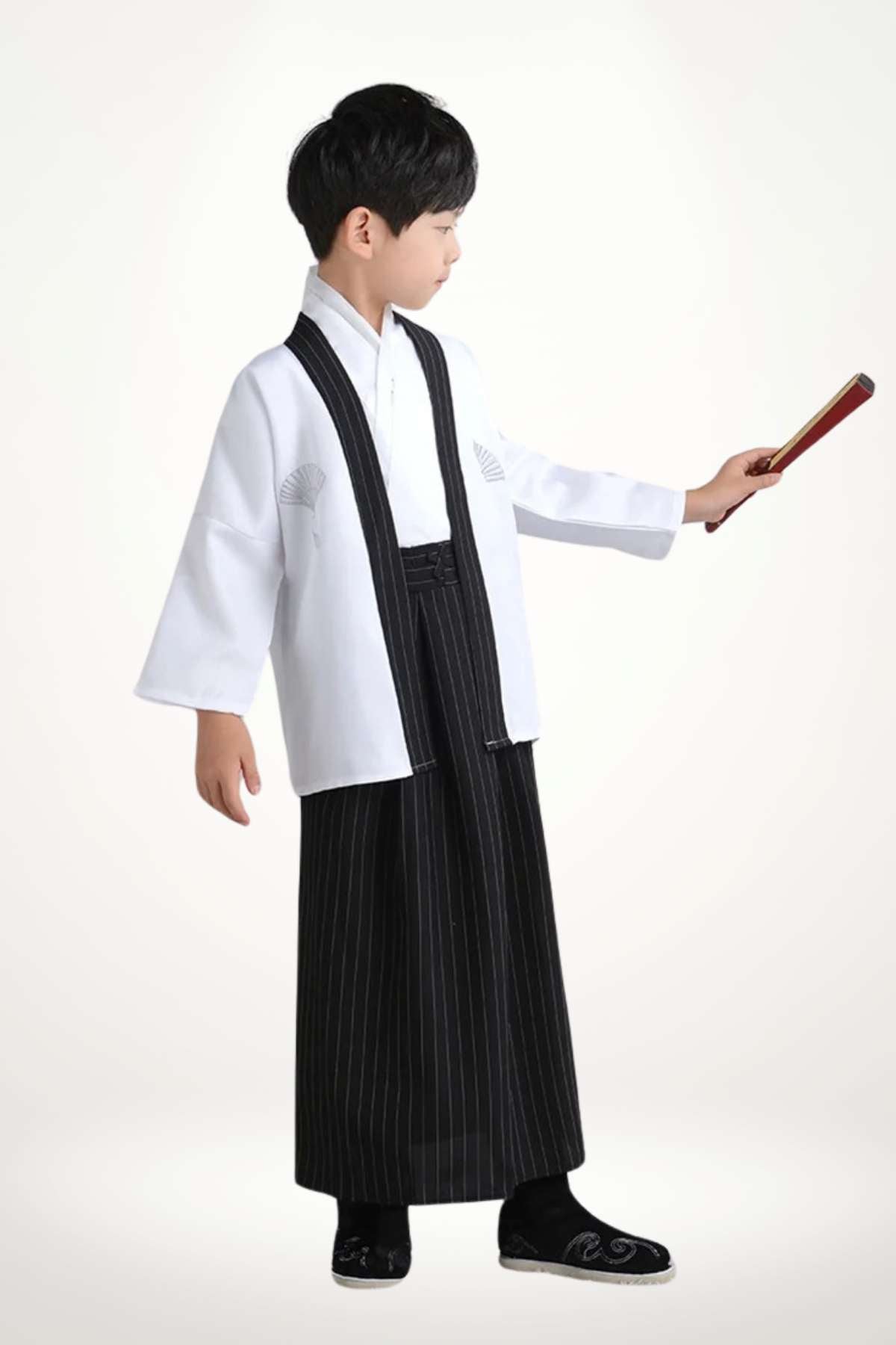 Enfant portant un kimono blanc avec des motifs, pantalon noir à rayures et chaussures noires, tenant un objet rouge