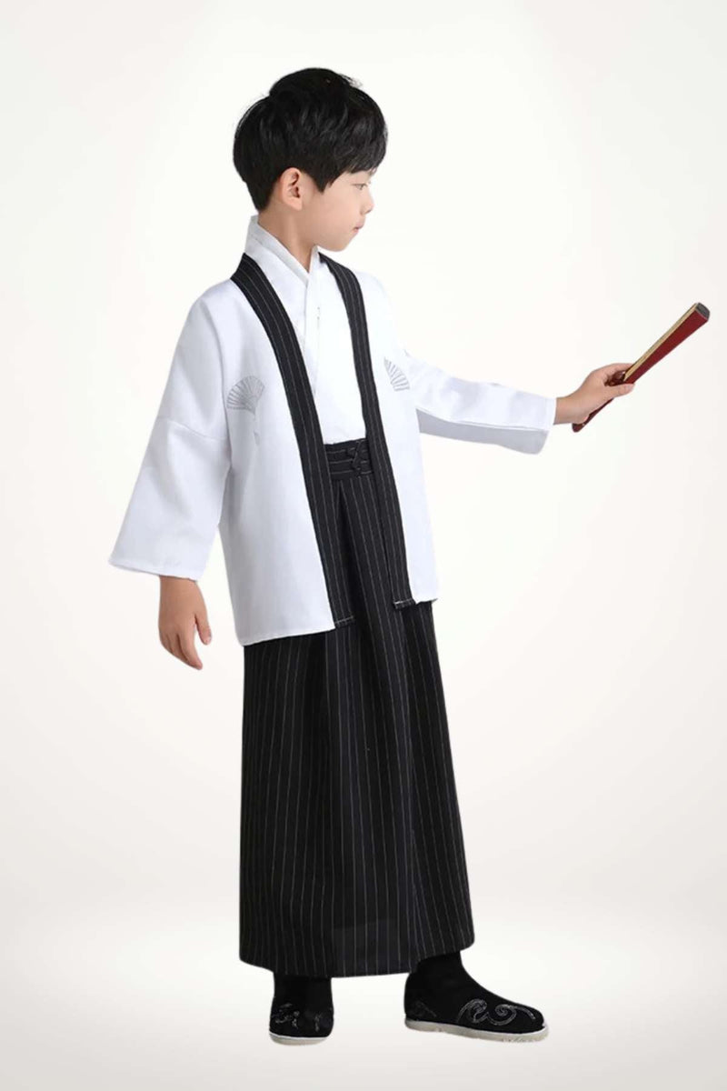 Enfant portant un kimono blanc avec des motifs, pantalon noir à rayures et chaussures noires, tenant un objet rouge