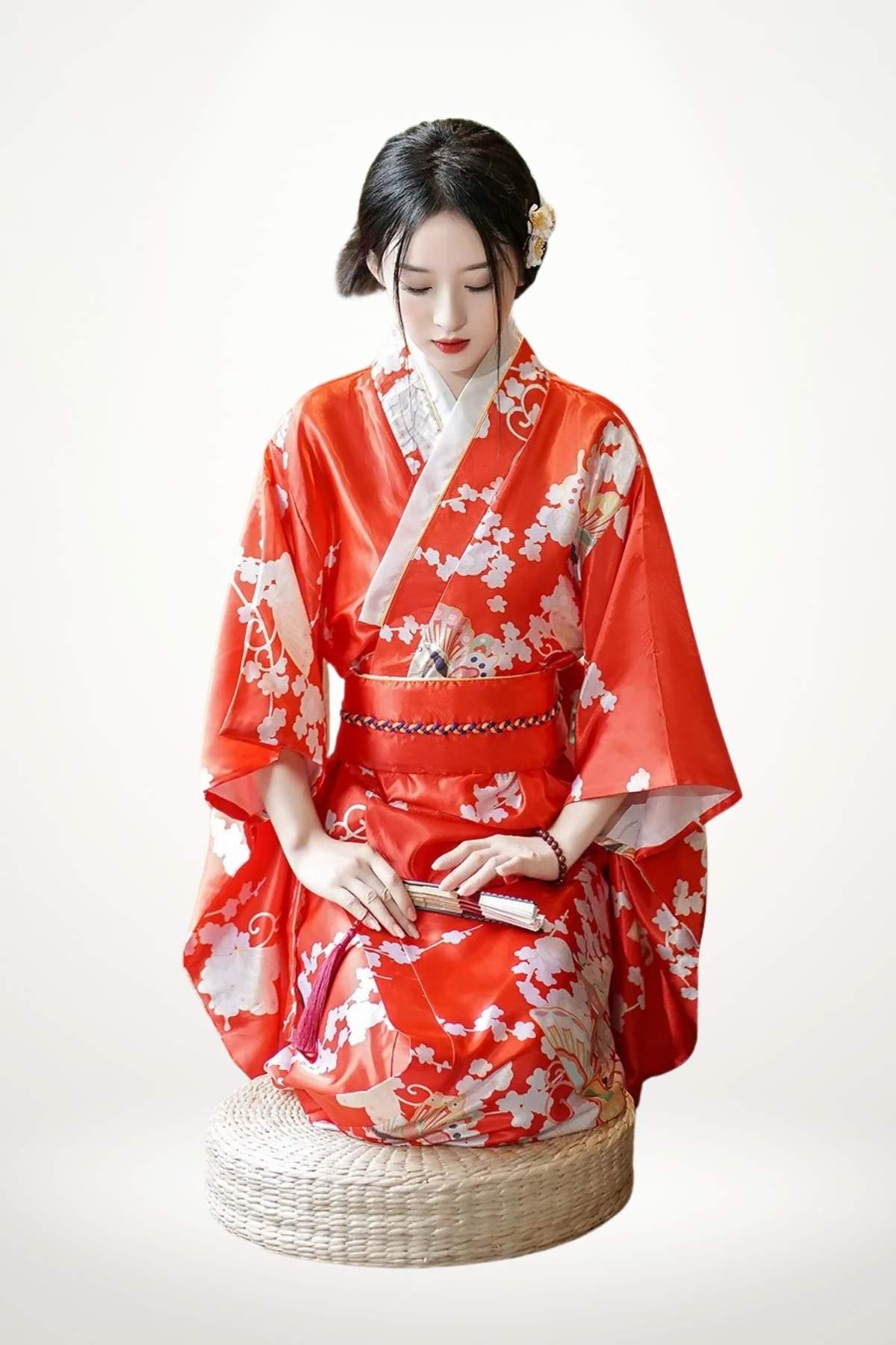Femme assise en kimono rouge orné de fleurs blanches, sur un coussin en osier, regard baissé