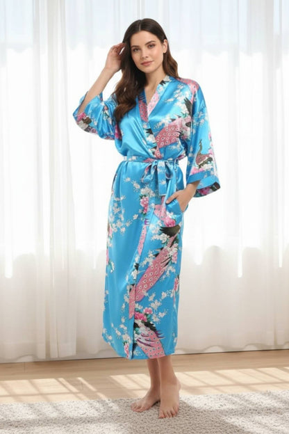 Femme en kimono bleu satiné orné de fleurs et oiseaux, posant dans une pièce lumineuse avec des rideaux blancs