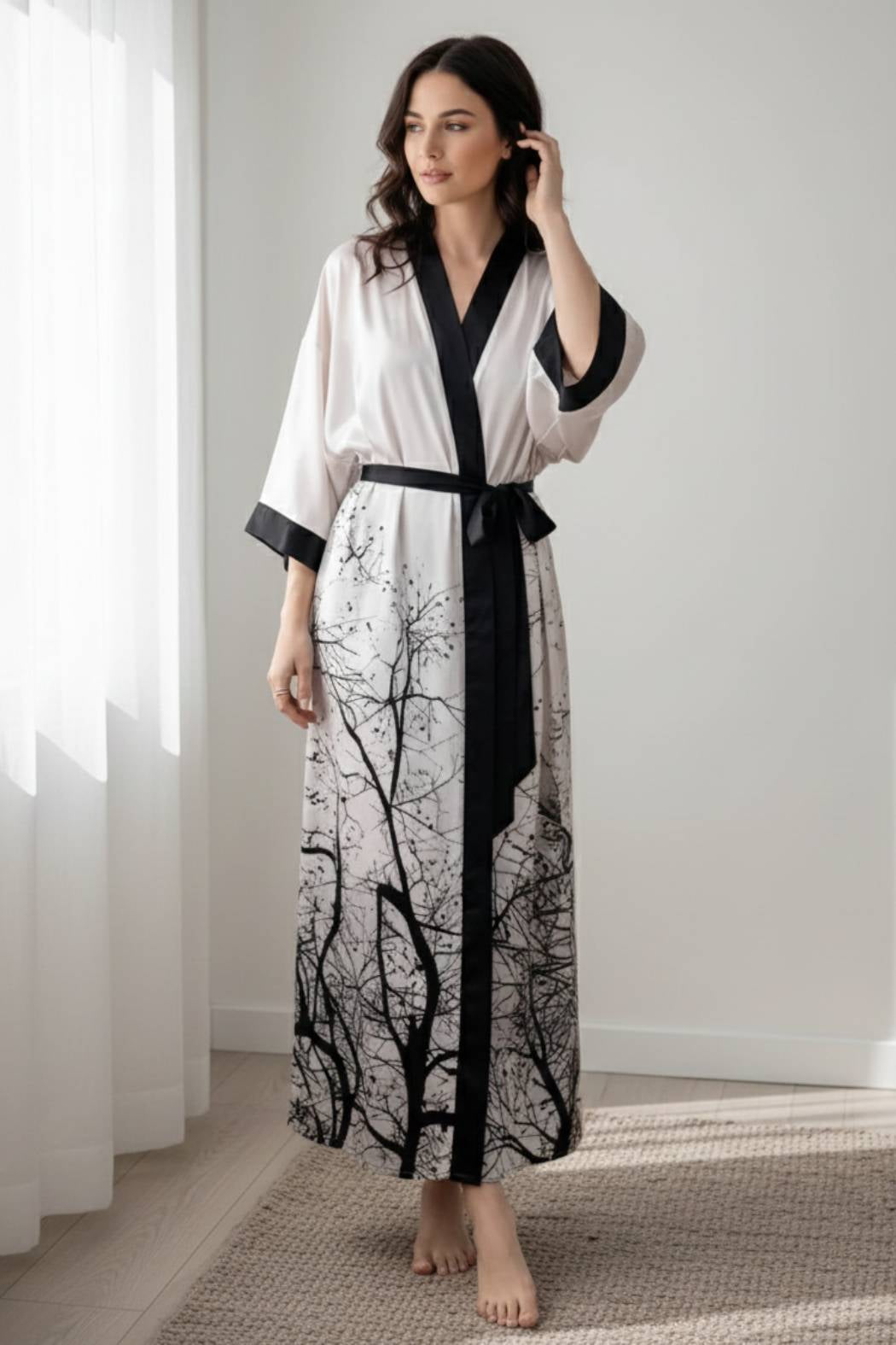 Femme en kimono long en satin blanc avec motifs noirs de branches, se tenant dans un décor lumineux et minimaliste