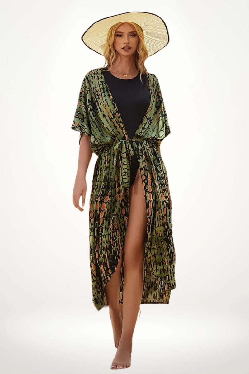 Femme en kimono vert à motifs, debout sur une plage, portant un chapeau large, avec une allure élégante