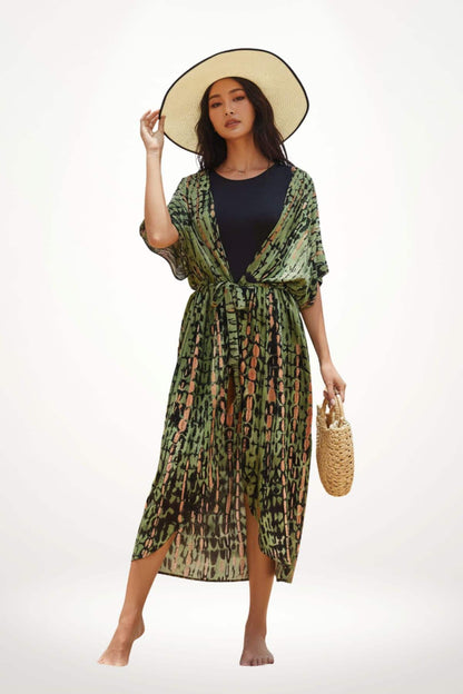 Femme posant en kimono vert à motifs, portant un chapeau large et tenant un sac en osier, sur fond neutre