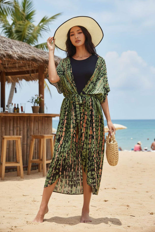 Femme portant un kimono vert à motifs sur une plage, chapeau large, décor tropical en arrière-plan