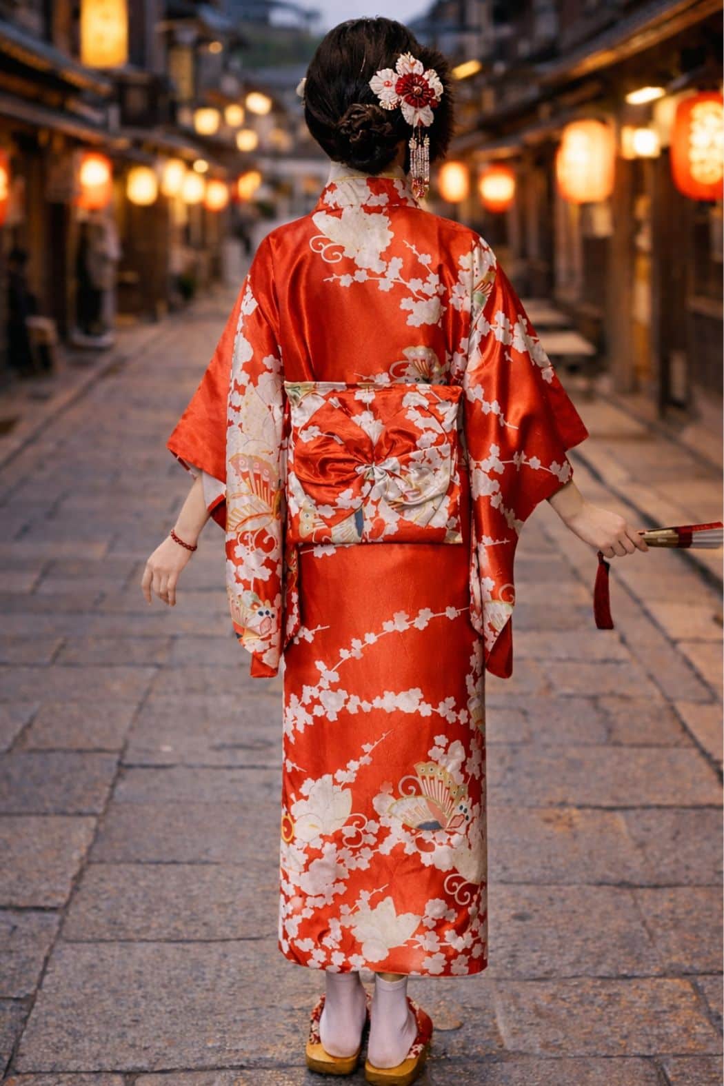 Femme en kimono rouge avec motifs floraux, se tenant dans une ruelle éclairée par des lanternes