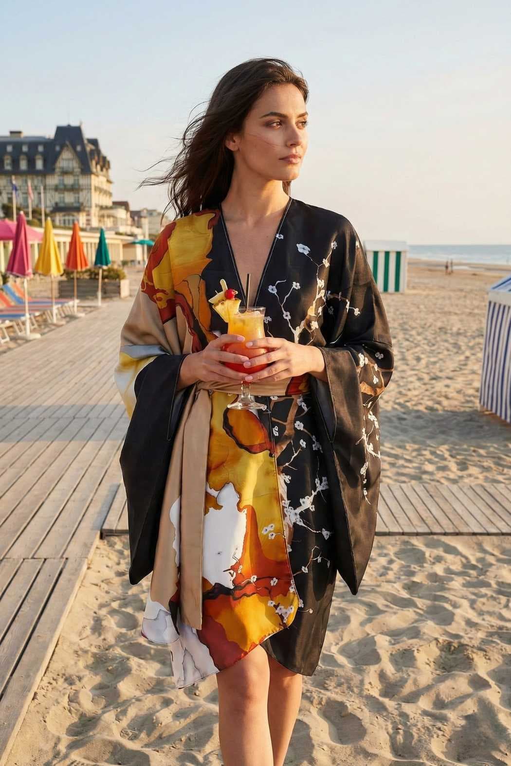 Femme portant un kimono satin aux motifs colorés, debout sur la plage avec un verre à la main