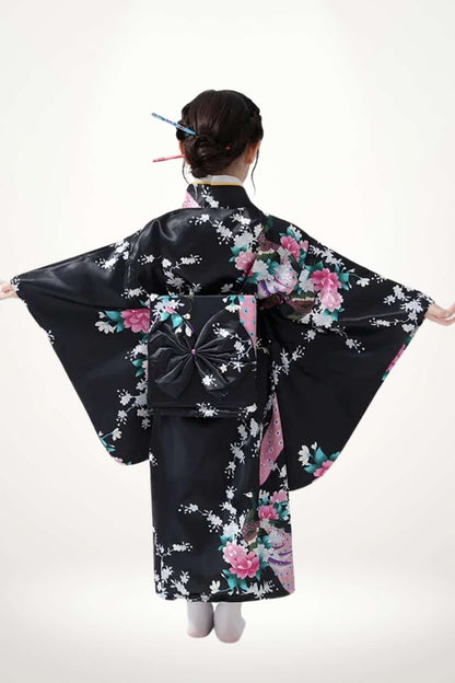 Fille en kimono noir avec motifs floraux colorés, bras écartés, décor sobre et élégant en arrière-plan