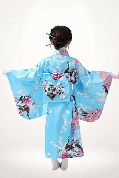Une jeune fille brune porte un kimono bleu orné de motifs floraux et de paons, avec un nœud décoratif dans le dos