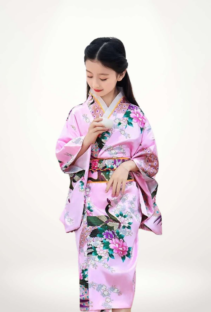 Une petite fille porte un kimono rose orné de fleurs et de motifs de paon, tenant une tasse