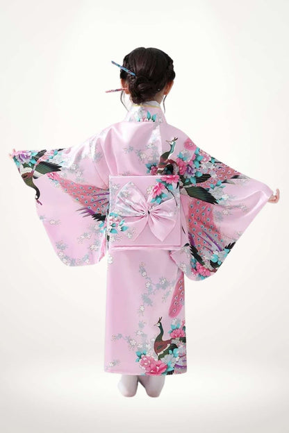 Une petite fille porte un kimono rose orné de motifs floraux et de paons, avec des manches larges et un nœud dans le dos