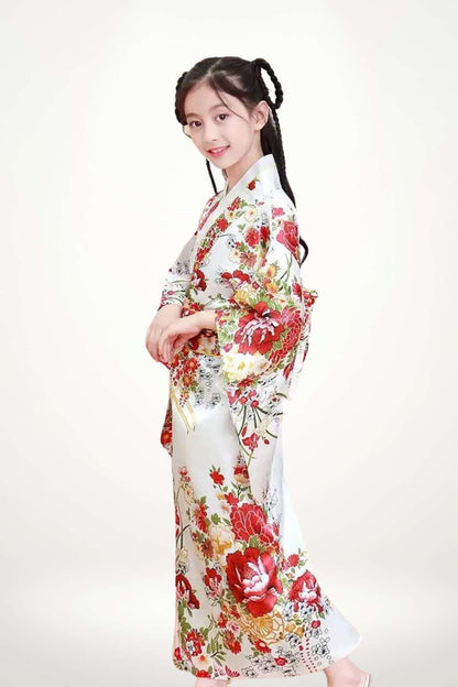 Une fillette souriante porte un kimono blanc orné de fleurs rouges et vertes, se tenant légèrement de profil