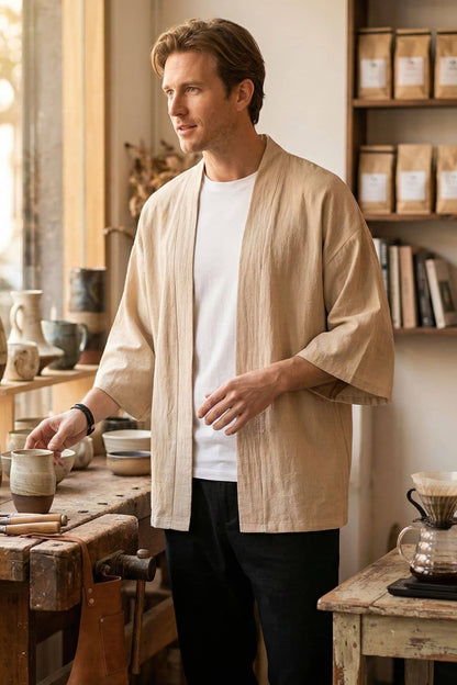 Homme en kimono beige, tenant une tasse, dans un décor chaleureux avec des poteries et des étagères en bois
