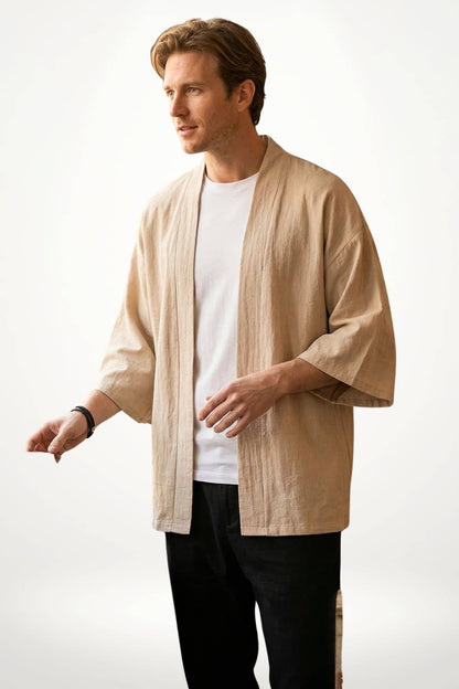 Homme en kimono beige, manches larges, se tenant debout sur fond blanc, avec un t-shirt blanc et un bracelet noir