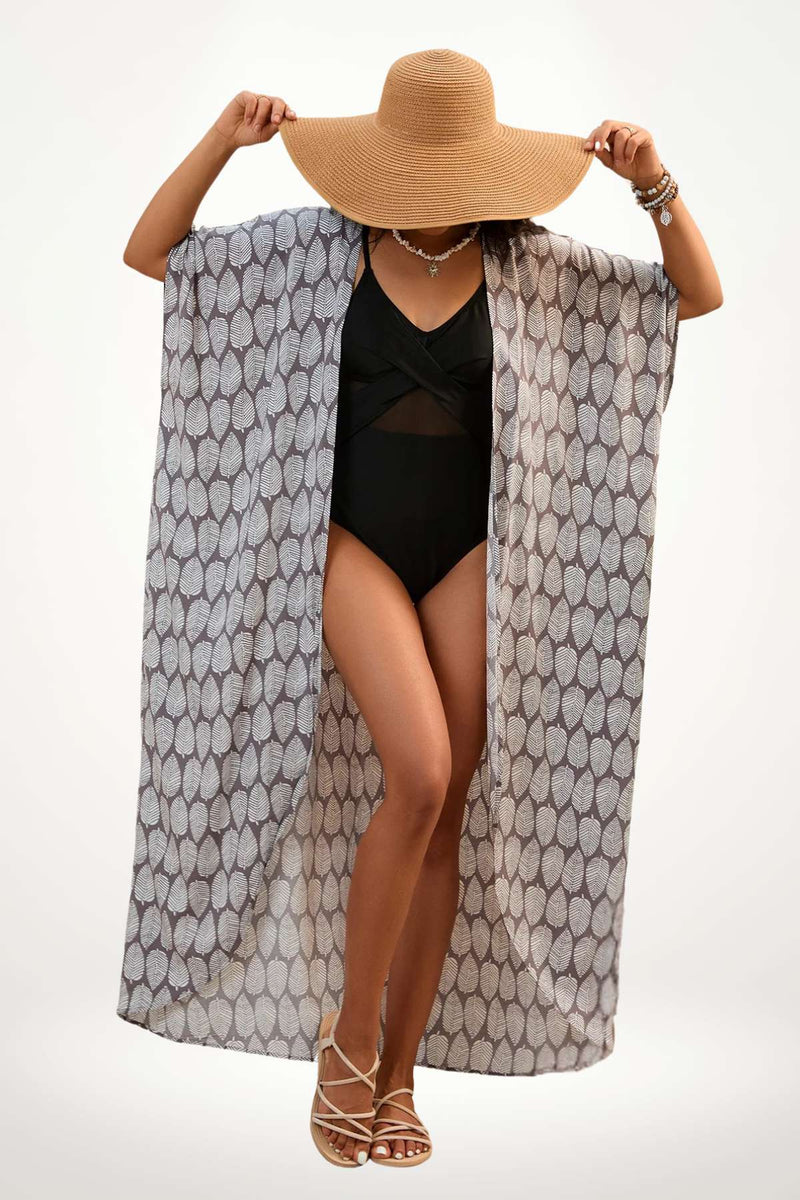 Femme portant un kimono léger à motifs de feuilles, chapeau large et maillot de bain noir, sur fond clair