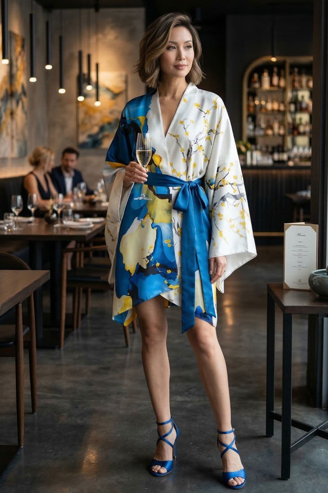 Femme en kimono satin blanc avec motifs bleus et jaunes, debout dans un restaurant élégant, tenant un verre